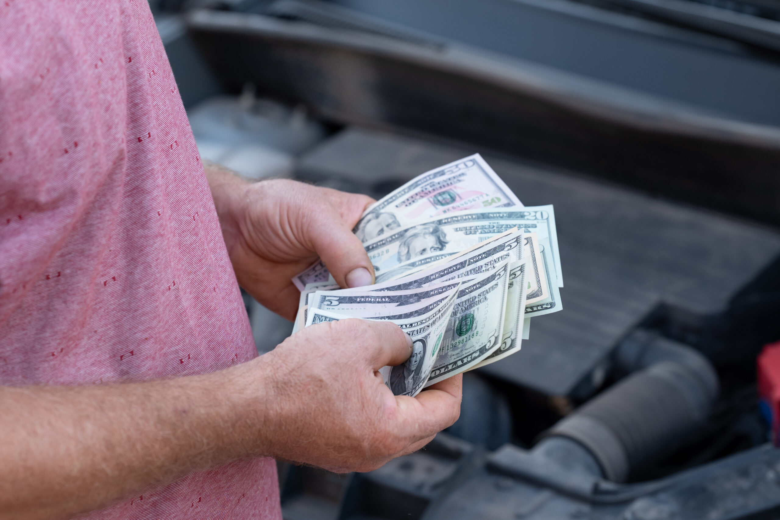 Holding Us Dollar Cash While Inspecting A Vehicle Under The Hood In A Parking Lot