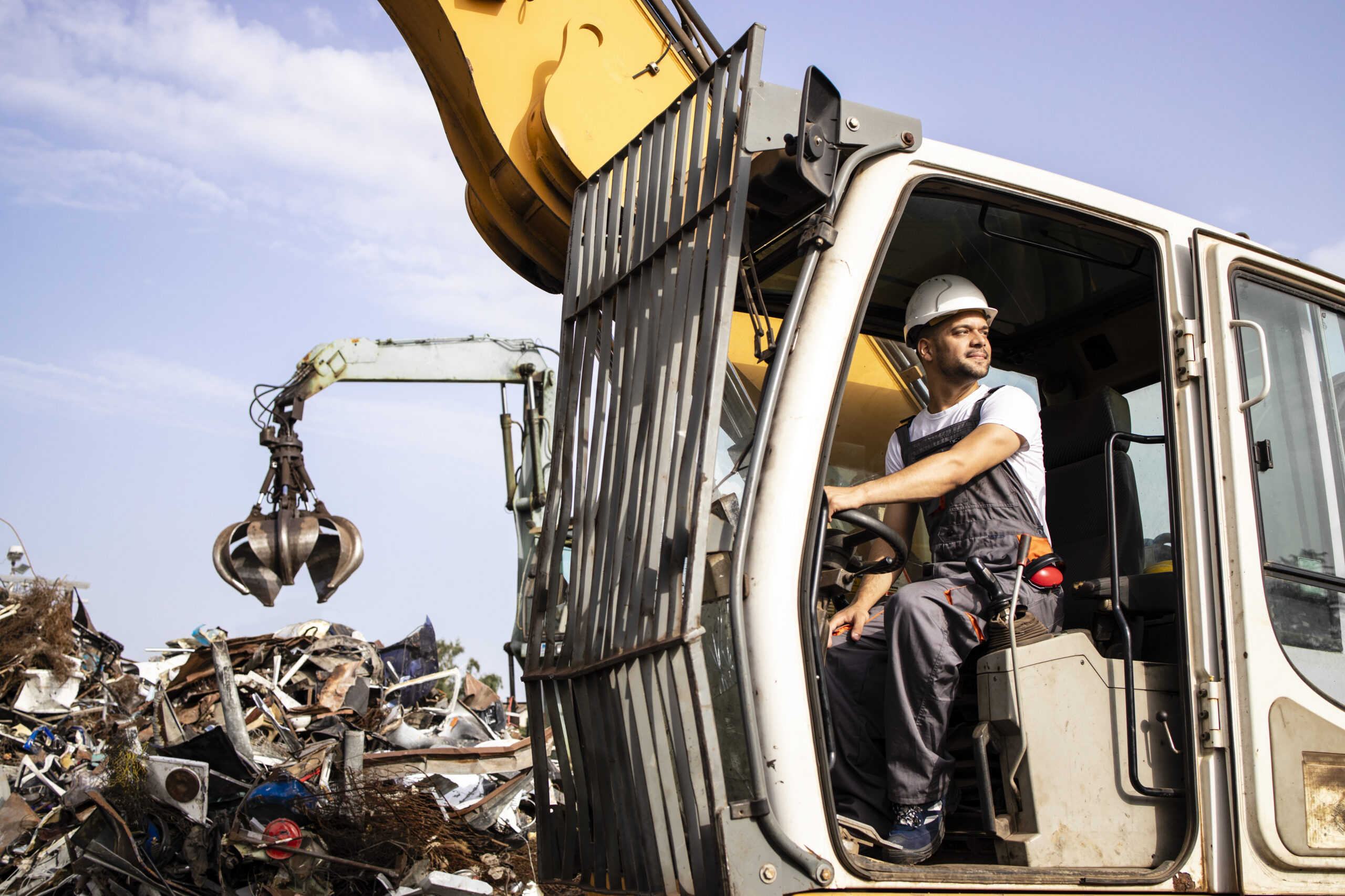 Man Operating Excavator Industrial Machine With Claw Attachment Used For Lifting Scrap Metal In Junk Yard.